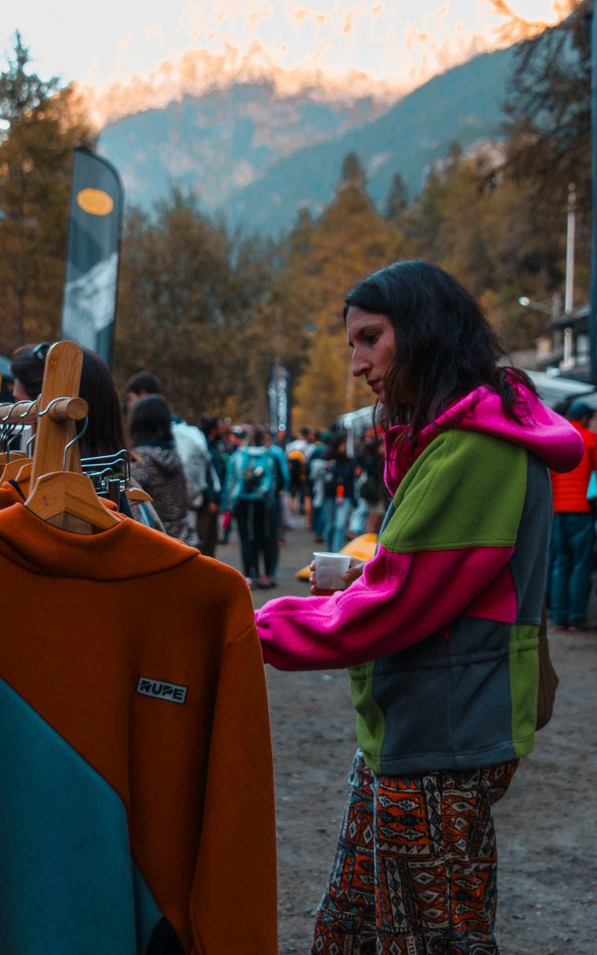 Una ragazza con un bicchiere di caffè guarda un espositore di felpe Rupe al villaggio del Valle Orco Climbing Festival, montagne al tramonto sullo sfondo